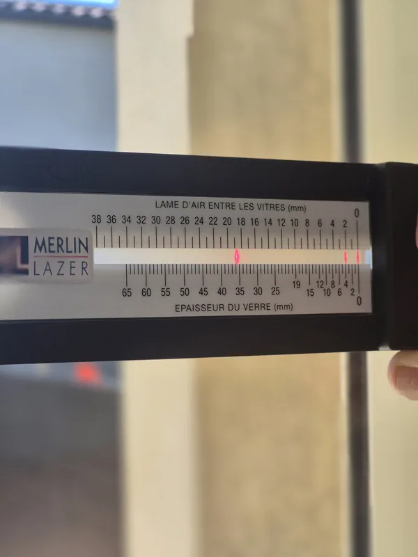 Calibre Merlin Lazer posé sur un double vitrage : deux réglettes graduées mesurent l’épaisseur du verre et la lame d’air entre les vitres.