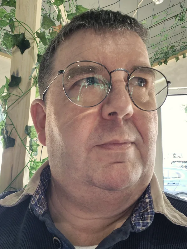 Portrait de Maurice Bremond, diagnostiqueur immobilier certifié, lunettes rondes et pull bleu marine, devant un treillage de lierre.
