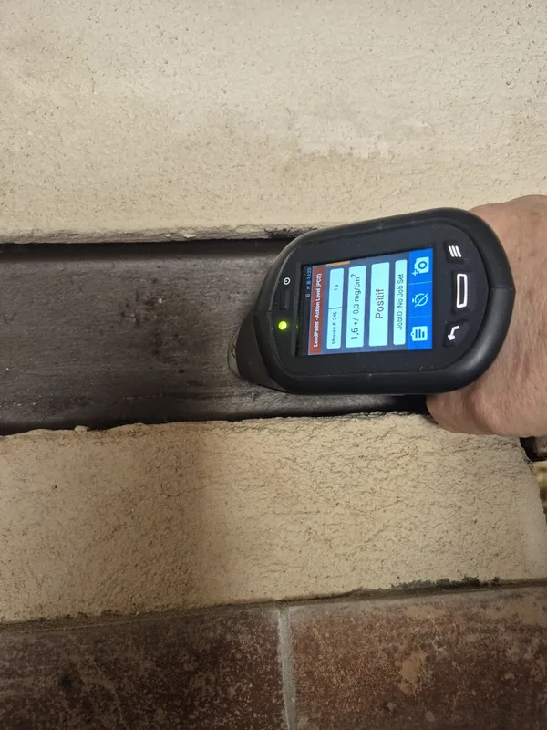 Analyseur de plomb portatif appliqué contre un dormant de fenêtre en bois dans un mur de pierre : l’écran affiche 1,6 ± 0,3 mg/cm² et le verdict « Positif ».
